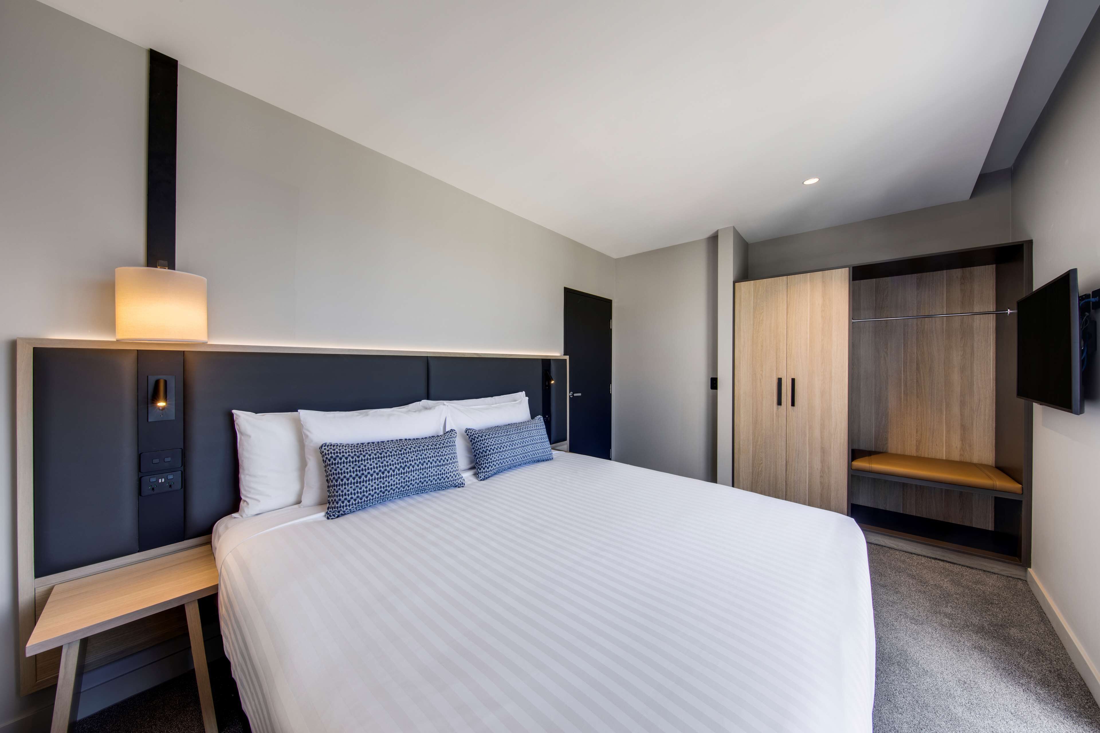 A modern hotel room with a large bed, bedside tables, a wardrobe, a television, and a built-in bench.