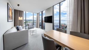 A modern hotel room with a large window, a flat-screen TV, a sofa, a dining table, and a small round table.