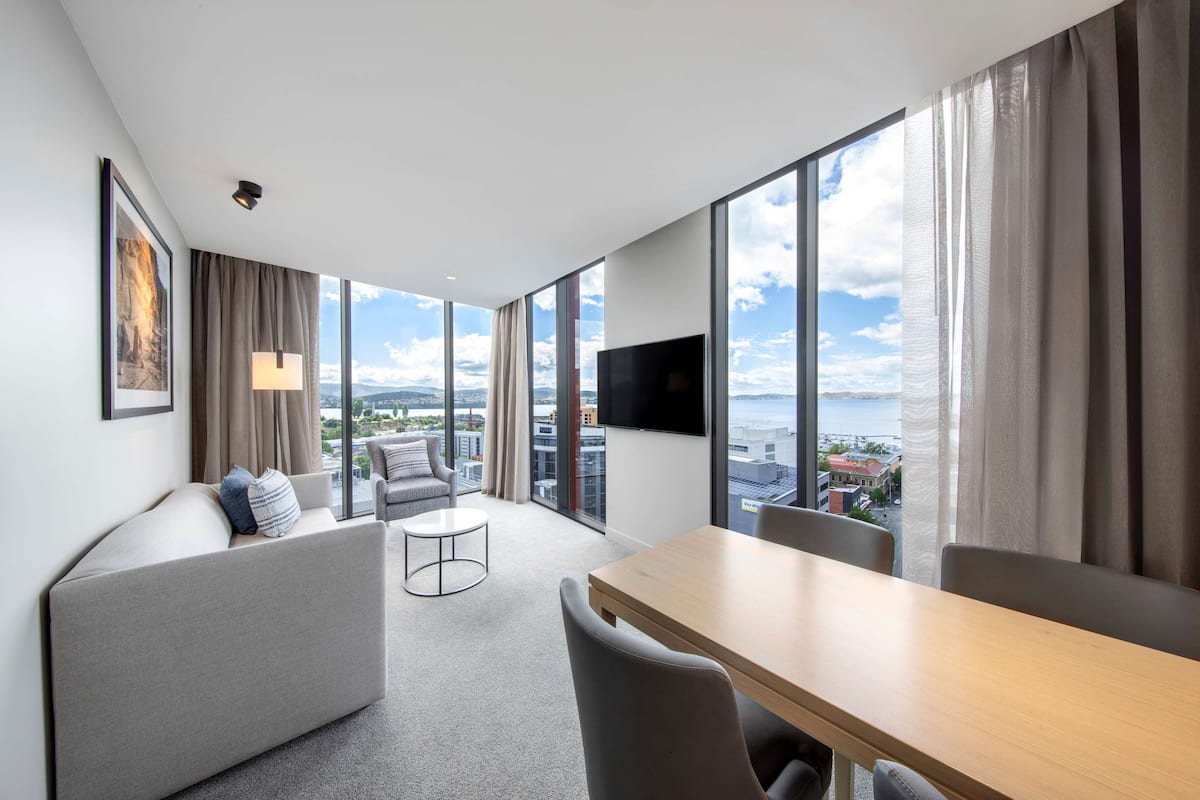Moderni hotellihuone, jossa on suuri ikkuna, taulu-tv, sohva, ruokapöytä ja pieni pyöreä pöytä.