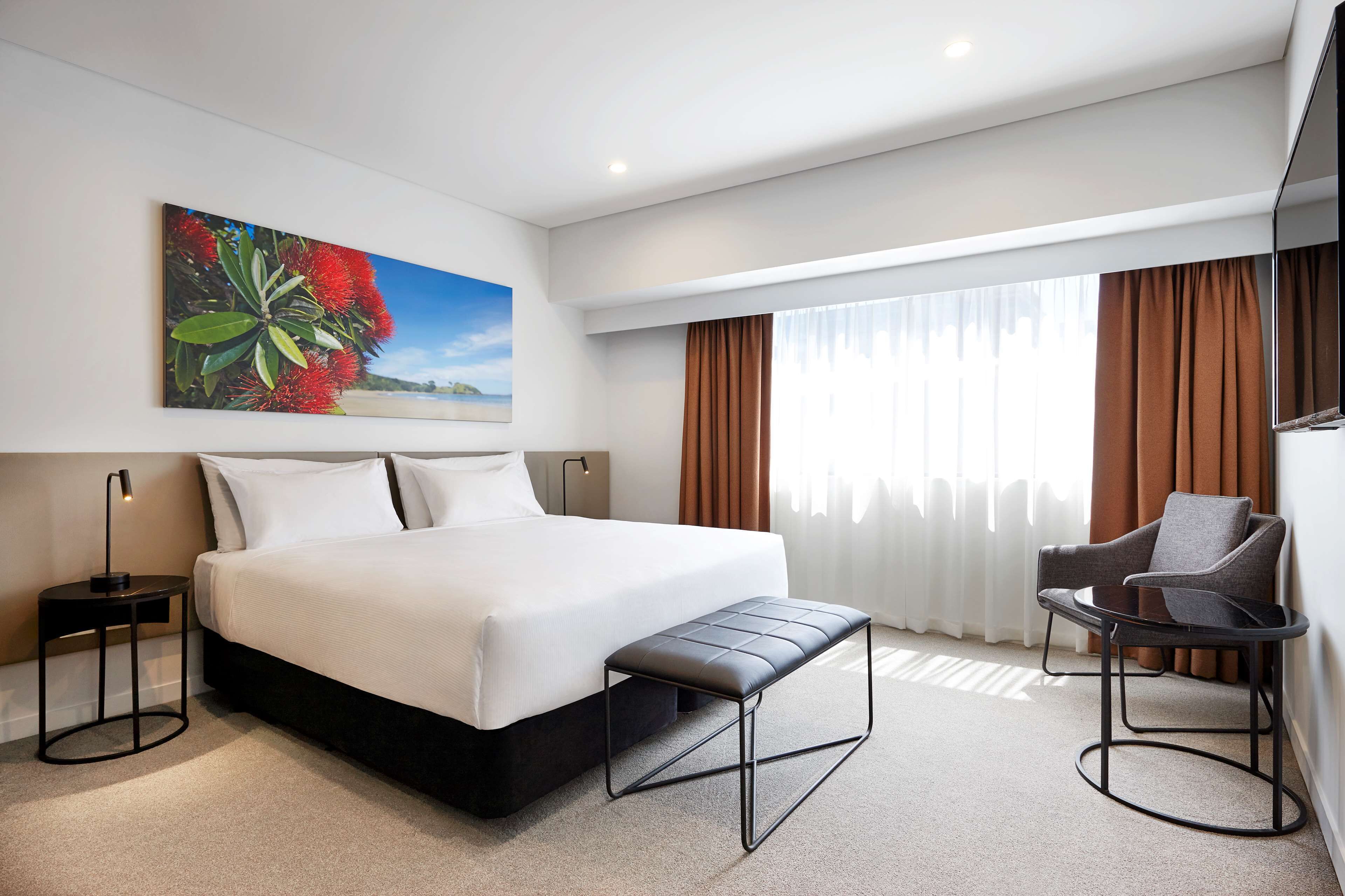 A modern hotel room with a large bed, a bench, a chair, a nightstand, and a painting of flowers.