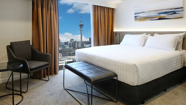 A hotel room with a large bed, a bench, a chair, and a view of a cityscape including a tower. - Travelodge Hotel Auckland Wynyard Quarter (Auckland)