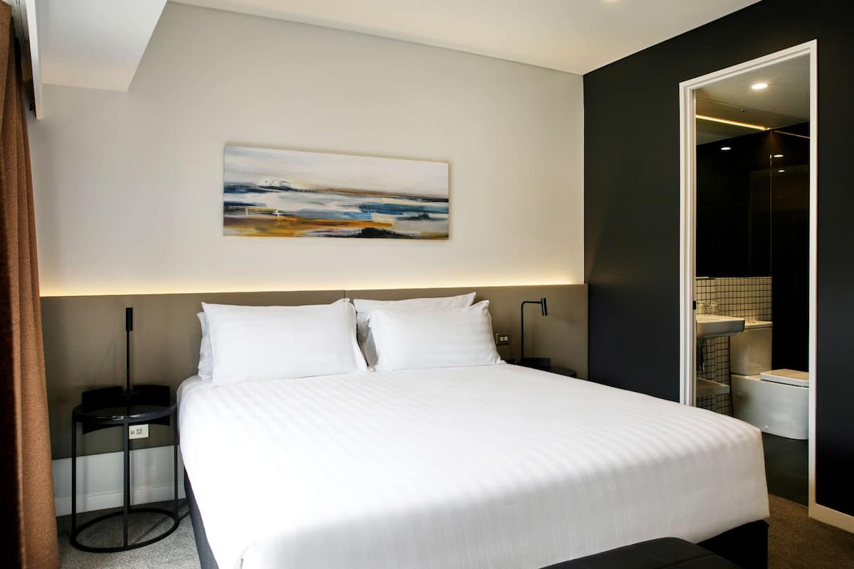 Moderni hotellihuone, jossa on suuri sänky, yöpöytä, seinälle ripustettu taulu ja kylpyhuone, joka näkyy avoimesta ovesta.