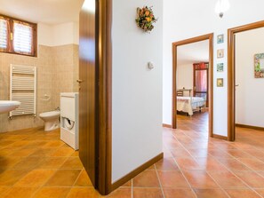 Ein Badezimmer mit Toilette, Bidet und Waschbecken. Eine Holztür führt in ein Schlafzimmer mit Bett und Fenster.
