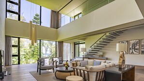 A spacious living area with a high ceiling, large windows offering a view of trees and a distant landscape, a staircase, and modern furniture. - UMITO OKINAWA ONNASON (Onna)