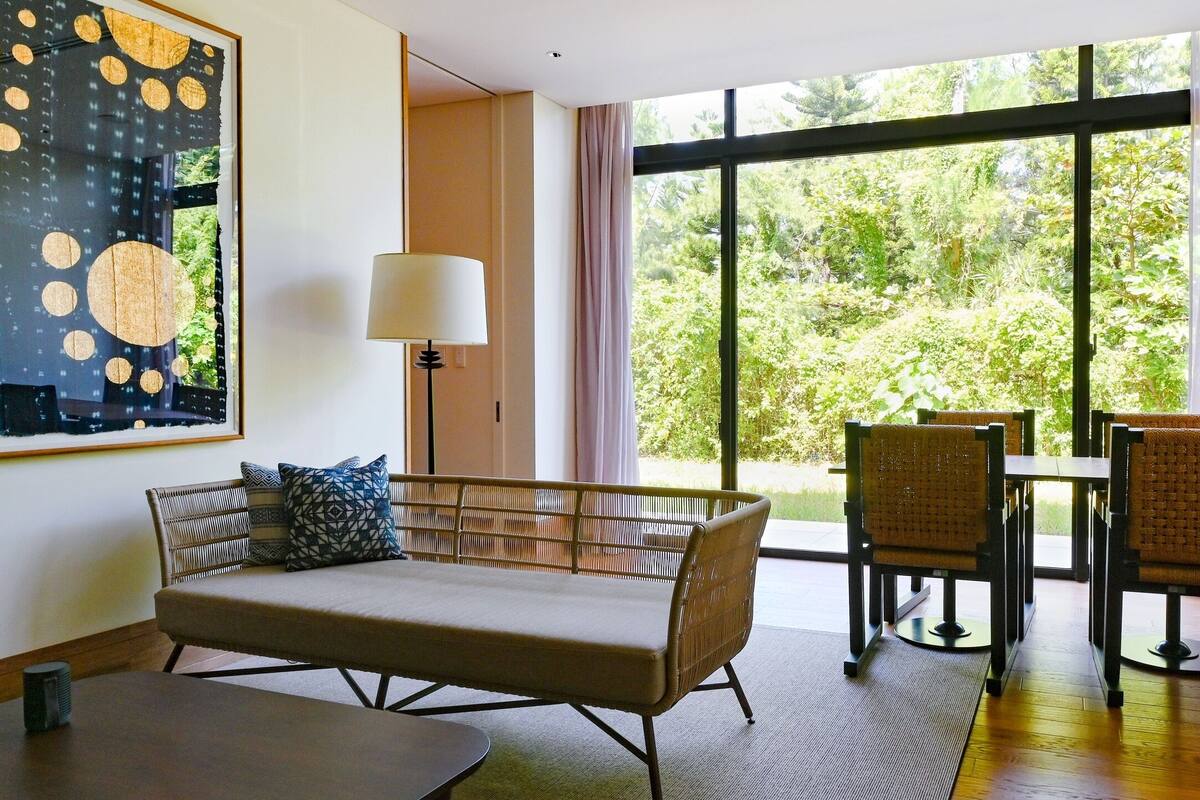 A modern room with a sofa, chairs, a table, and a large window offering a view of greenery.