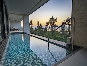 A modern indoor swimming pool with a clear view of trees and a sunset. - UMITO OKINAWA ONNASON (Onna)