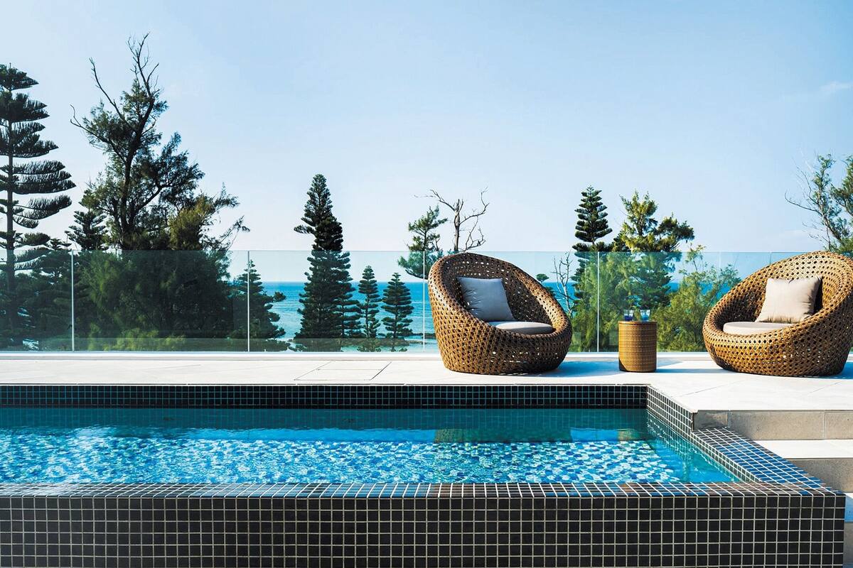 A swimming pool with two wicker chairs and a view of the ocean.