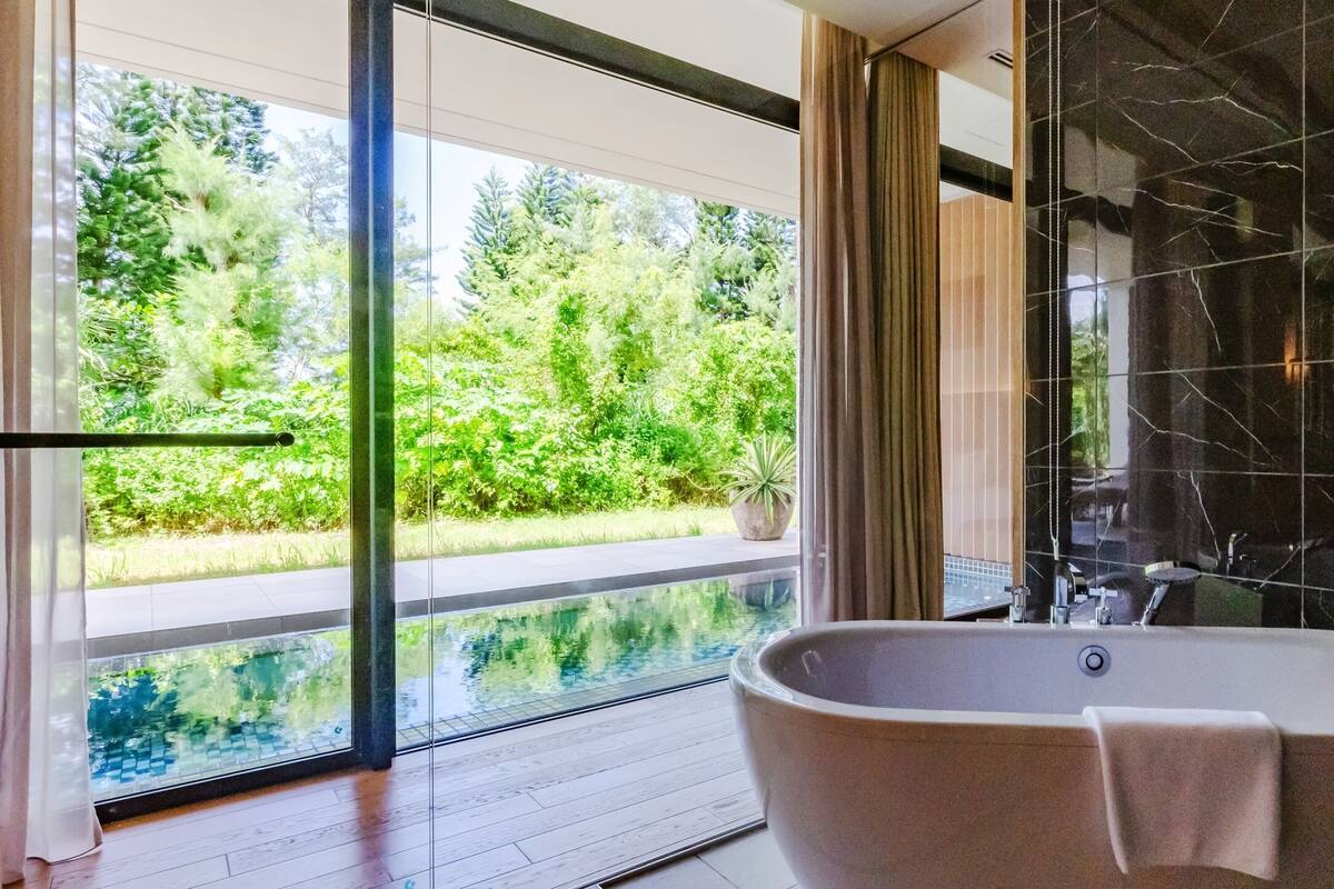 A modern bathroom with a freestanding bathtub, a glass-enclosed shower area, and a view of lush greenery outside.