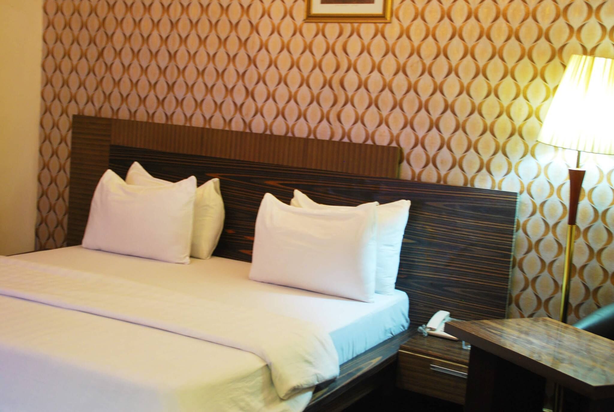 Una cama bien hecha con sábanas y almohadas blancas, cabecera de madera, mesita de noche con lámpara y pared con estampado.