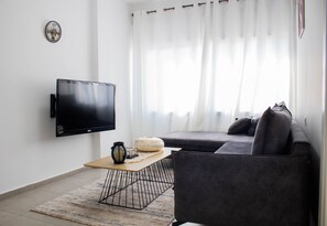 A modern living room with a flat-screen TV, a grey sofa, a coffee table, and a large window with curtains.