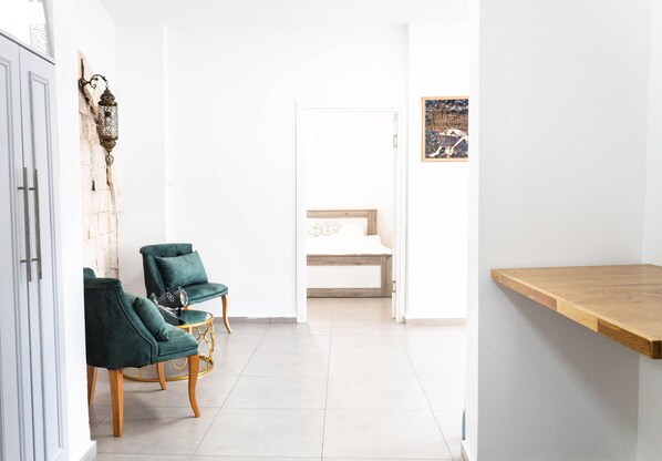 A hallway with a bed, two green chairs, a wooden table, and a wall-mounted lamp.