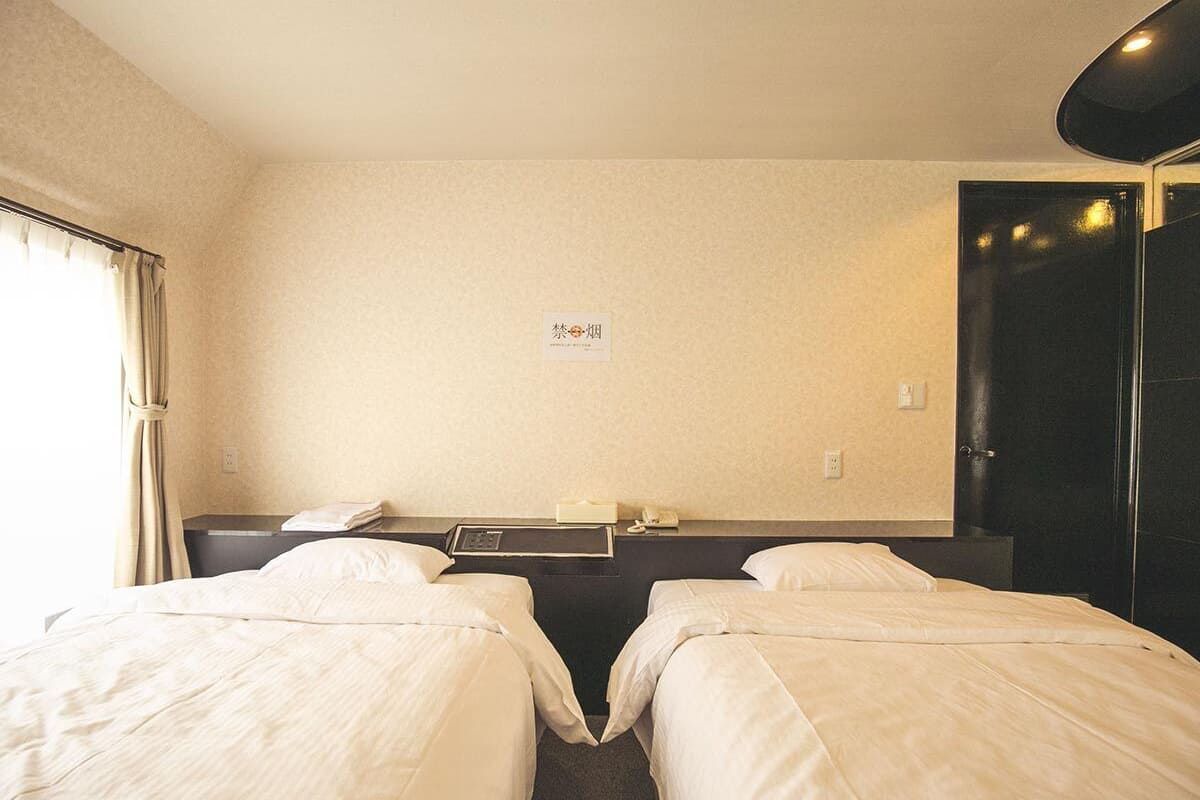 A hotel room with two single beds, a bedside table with a lamp, a small table with a phone and a book, and a wall-mounted sign.