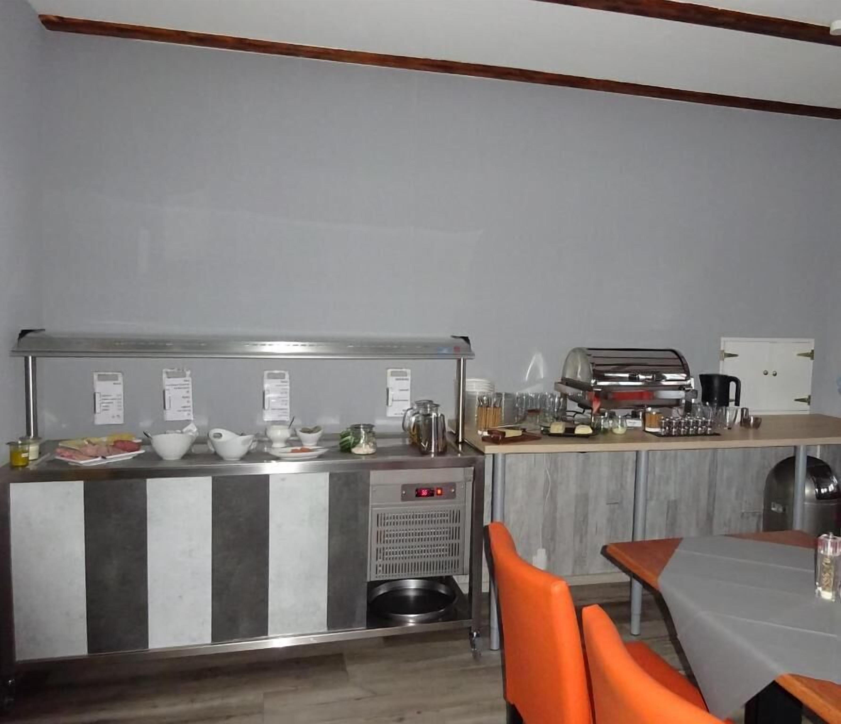 Una kitchenette con cafetera, hervidor de agua y diferentes utensilios de cocina. Hay un pequeño cubo de basura y una zona de estar con sillas anaranjadas.