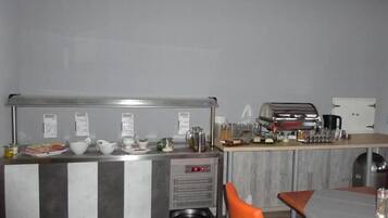 Una kitchenette con cafetera, hervidor de agua y diferentes utensilios de cocina. Hay un pequeño cubo de basura y una zona de estar con sillas anaranjadas.
