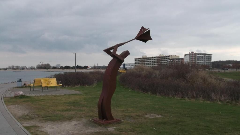 A sculpture of a figure holding a large object, located near a pathway and a body of water, with buildings in the background.