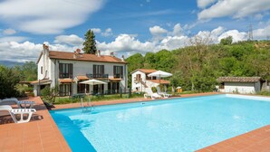 Une piscine entourée d’une terrasse carrelée et bordée de deux bâtiments blancs aux toits recouverts de tuiles rouges, dont l’un possède un balcon.