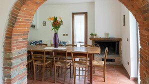 Un coin repas agrémenté d’une arche en briques, d’une table en bois et de chaises. On y trouve un vase avec des fleurs sur la table, une cheminée et une fenêtre avec des rideaux.
