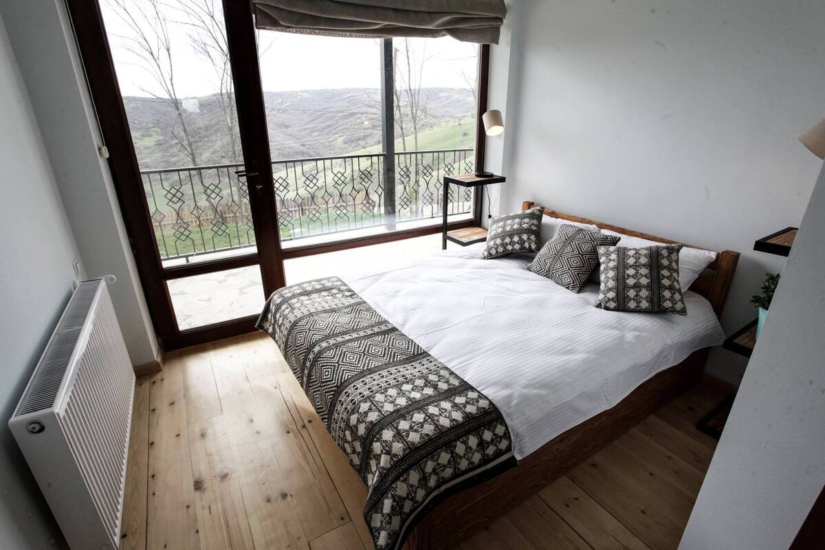 A bedroom with a large window, a bed with patterned bedding, a bedside table, and a radiator.