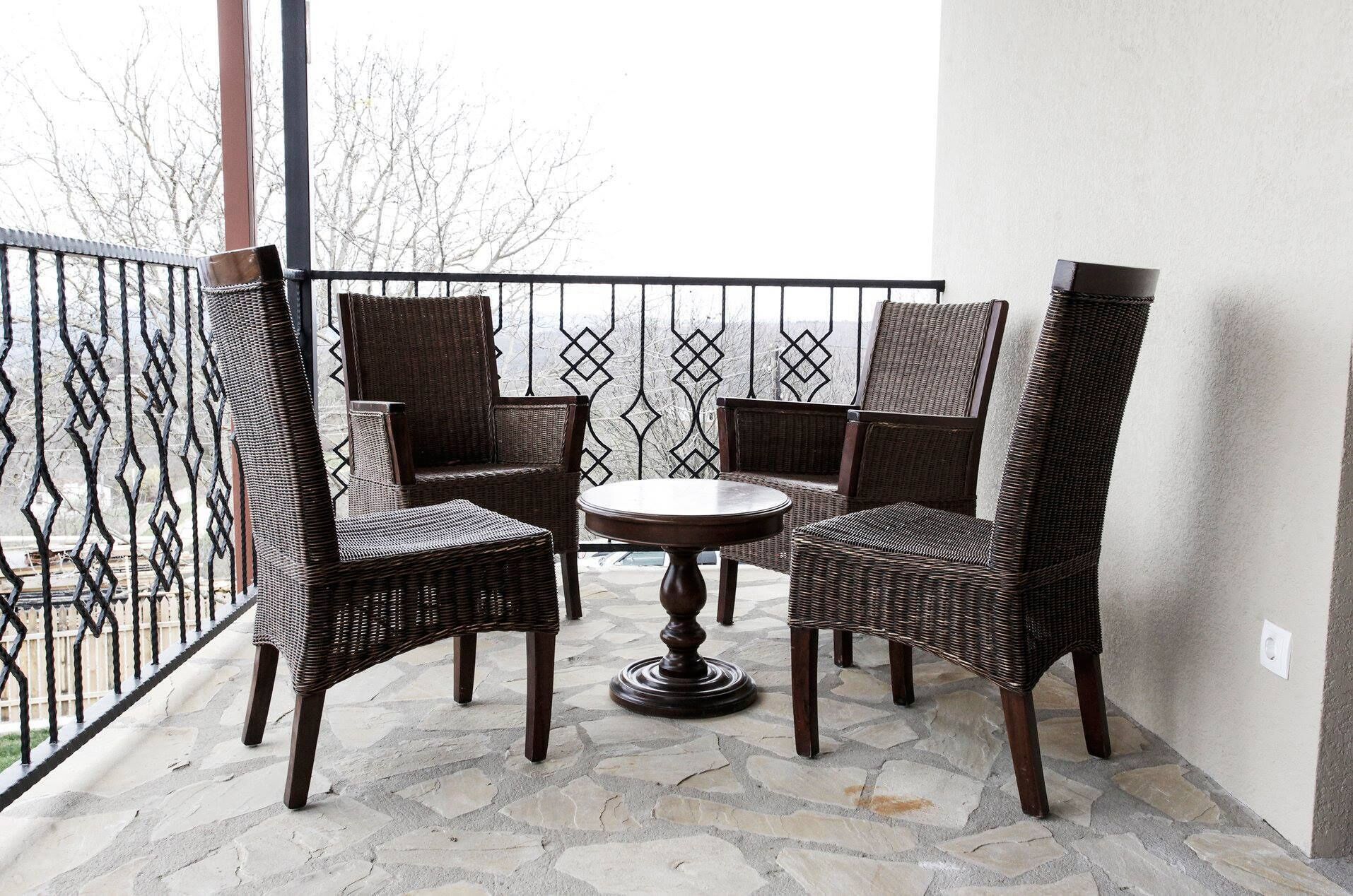 A balcony with wicker chairs and a round table, featuring a stone floor and a metal railing.