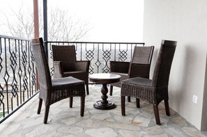 A balcony with wicker chairs and a round table, featuring a stone floor and a metal railing.