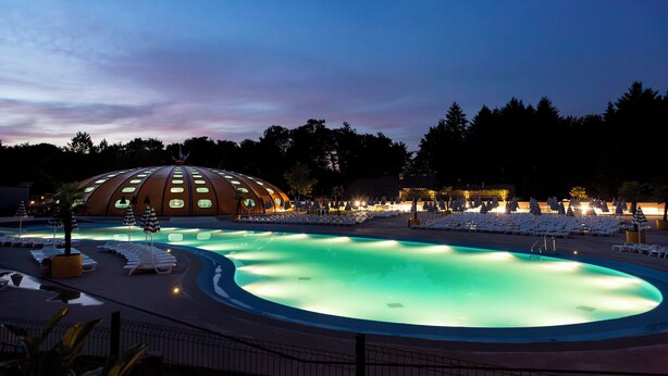 A swimming pool with lounge chairs and umbrellas, surrounded by a dome-shaped building with circular windows. - Hu Birkelt Village (Larochette)