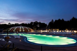 A swimming pool with lounge chairs and umbrellas, surrounded by a dome-shaped building with circular windows. - Hu Birkelt Village (Larochette)