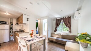 A modern kitchen with white cabinets, a microwave, and a refrigerator. There is a dining area with a white table and chairs, and a sofa with a window view. - Hu Birkelt Village (Larochette)