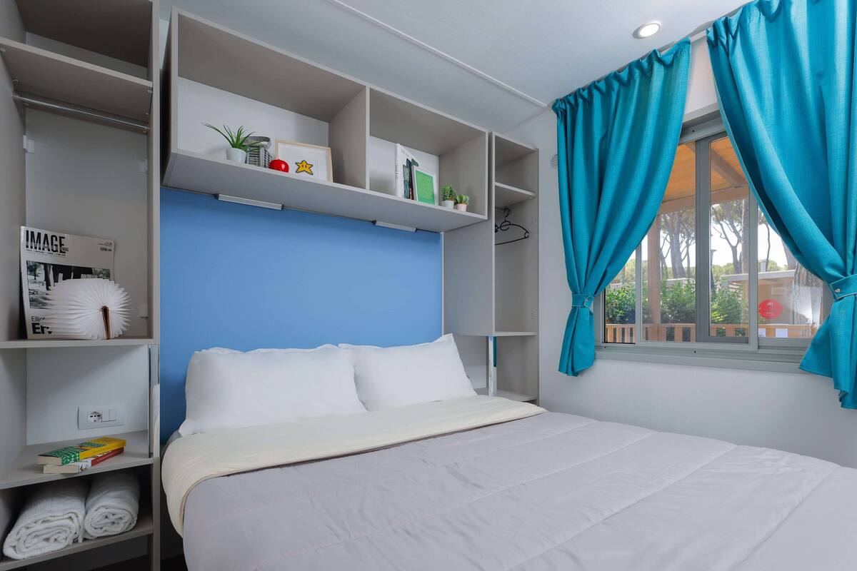 A neatly organized bedroom with a bed, white pillows, a blue accent wall, and shelves with various items.
