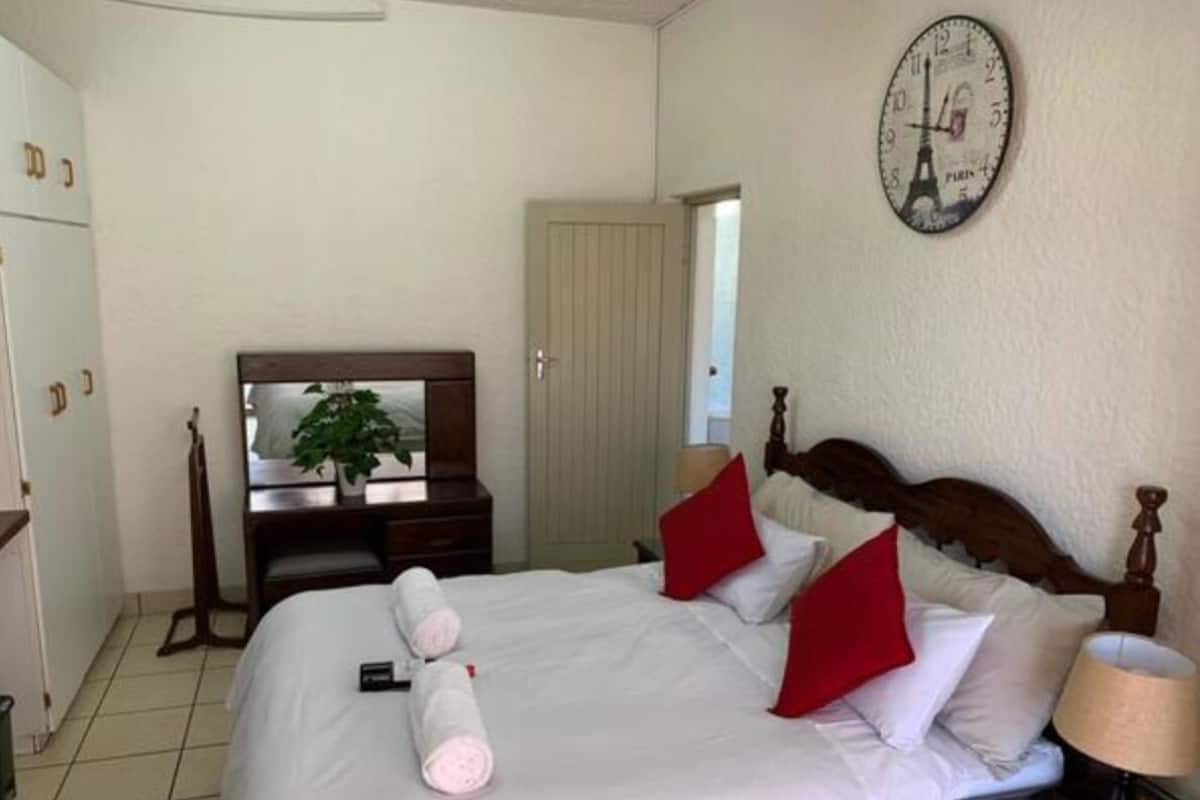 Una habitación de hotel con una cama, cabecera de madera, un reloj, cómoda y una planta.
