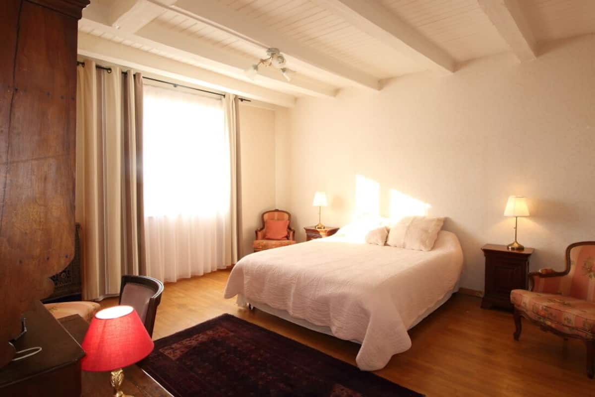 A bedroom with a large bed, two bedside lamps, a chair, a desk, and a window with curtains.