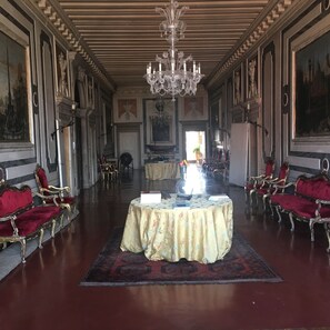 A grand room with ornate chandeliers, red upholstered chairs, and a central table with a floral tablecloth.