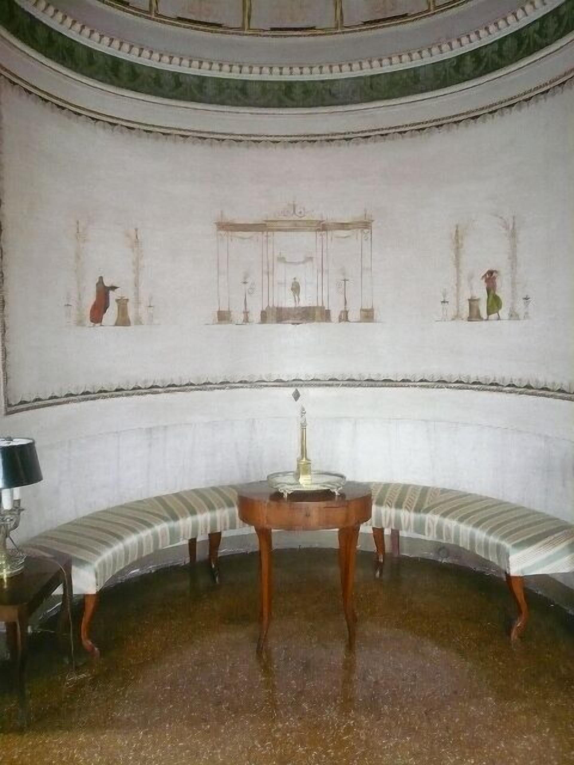 A room with a curved bench, a small table with a lamp, and a wall with classical-style paintings.