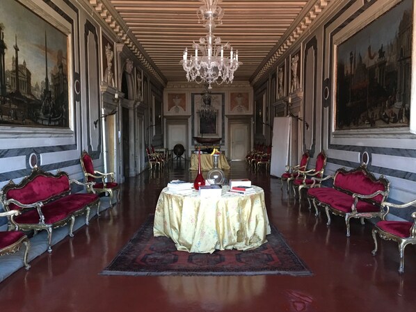 A grand room with ornate chandeliers, red velvet chairs, and a central table with books and a vase.