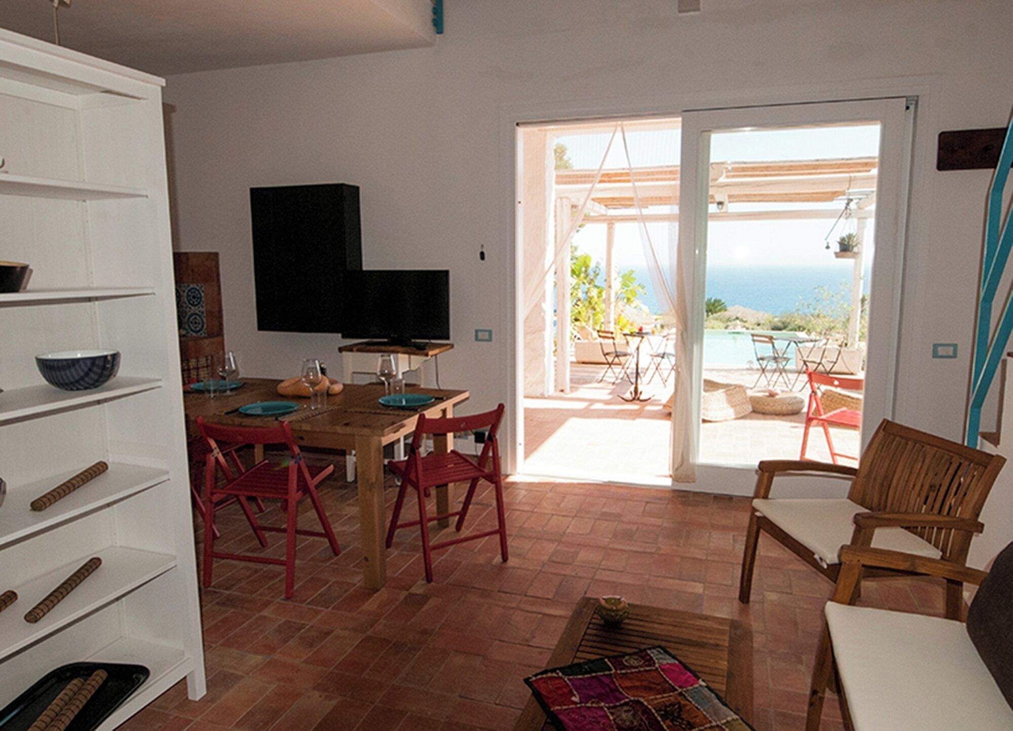 A dining area with a wooden table and red chairs, a television on the wall, and a view of the sea through the open door.