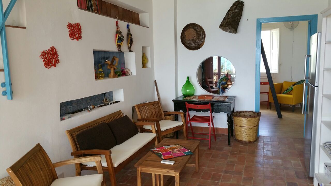 Uma sala de estar com sofá, cadeiras, uma mesa e uma área de refeições com uma cadeira e uma mesa pequena. Há itens decorativos nas paredes e um espelho sobre a mesa de jantar.