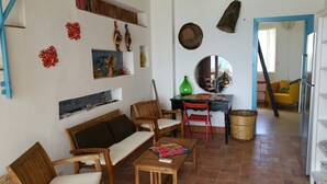 Ein Wohnzimmer mit einer Couch, Sesseln, einem Tisch und einem Essbereich mit einem Stuhl und einem kleinen Tisch. An den Wänden befinden sich dekorative Elemente und ein Spiegel über dem Esstisch.