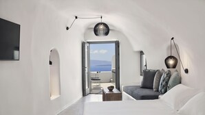 Un dormitorio moderno y minimalista con cama, zona de estar con cojines, una mesita y vistas al mar a través de una puerta abierta.