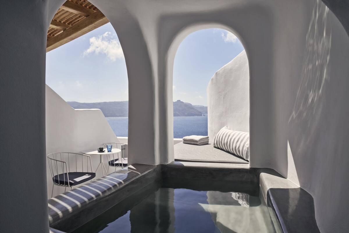 Una habitación moderna y minimalista con vistas despejadas al mar y montañas distantes. Cuenta con una zona de estar con un banco acolchado y una pequeña mesa con una taza.