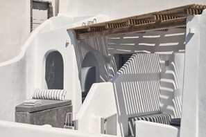 A white-walled room with a wooden pergola, a cushioned seating area, and a small table with a towel. - Alisaris Caves (Santorini)