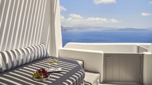 Un balcón con vista al mar, una mesa con un cuenco de uvas y copas de vino, y un cojín a rayas.