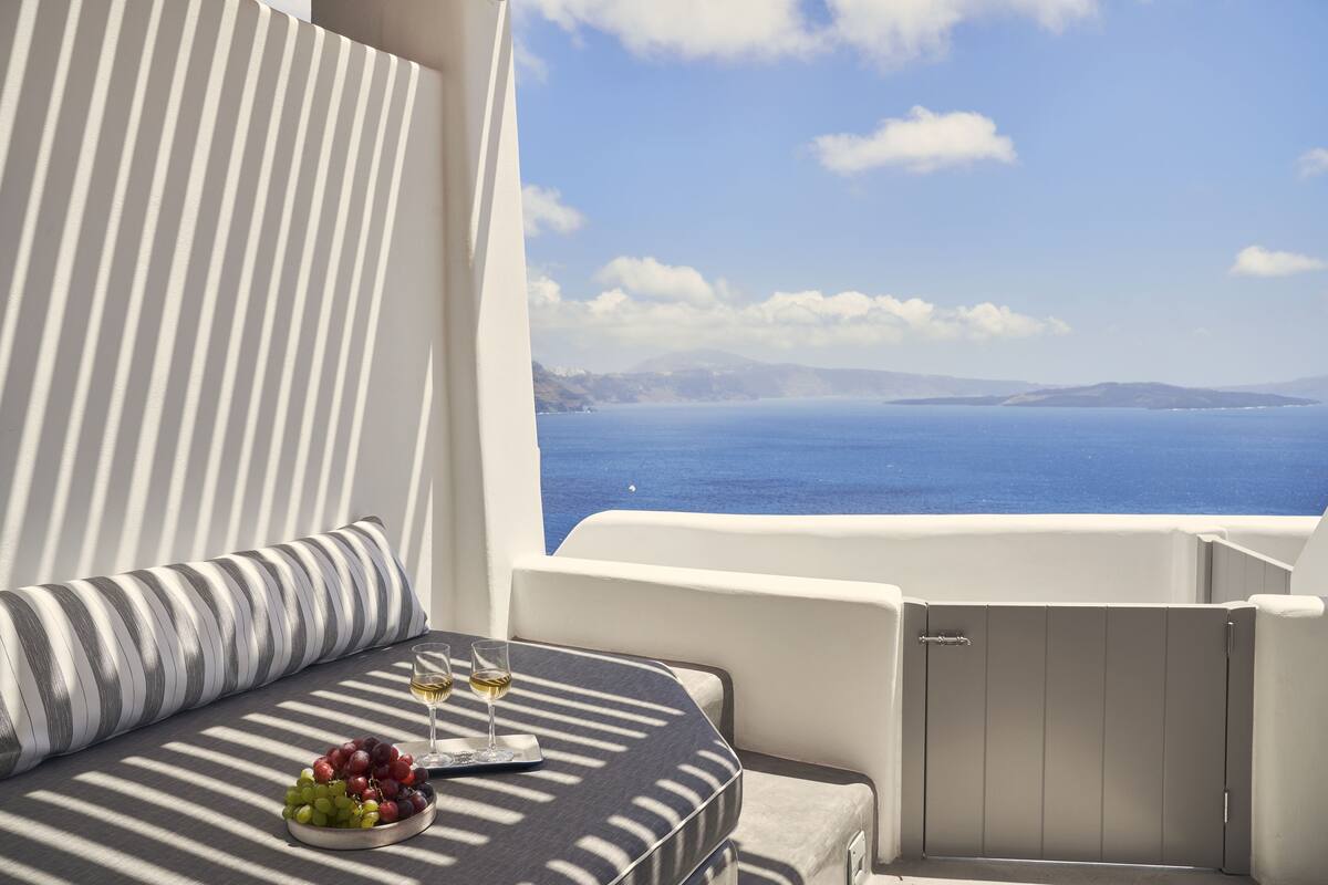 Un balcón con vista al mar, una mesa con un cuenco de uvas y copas de vino, y un cojín a rayas.