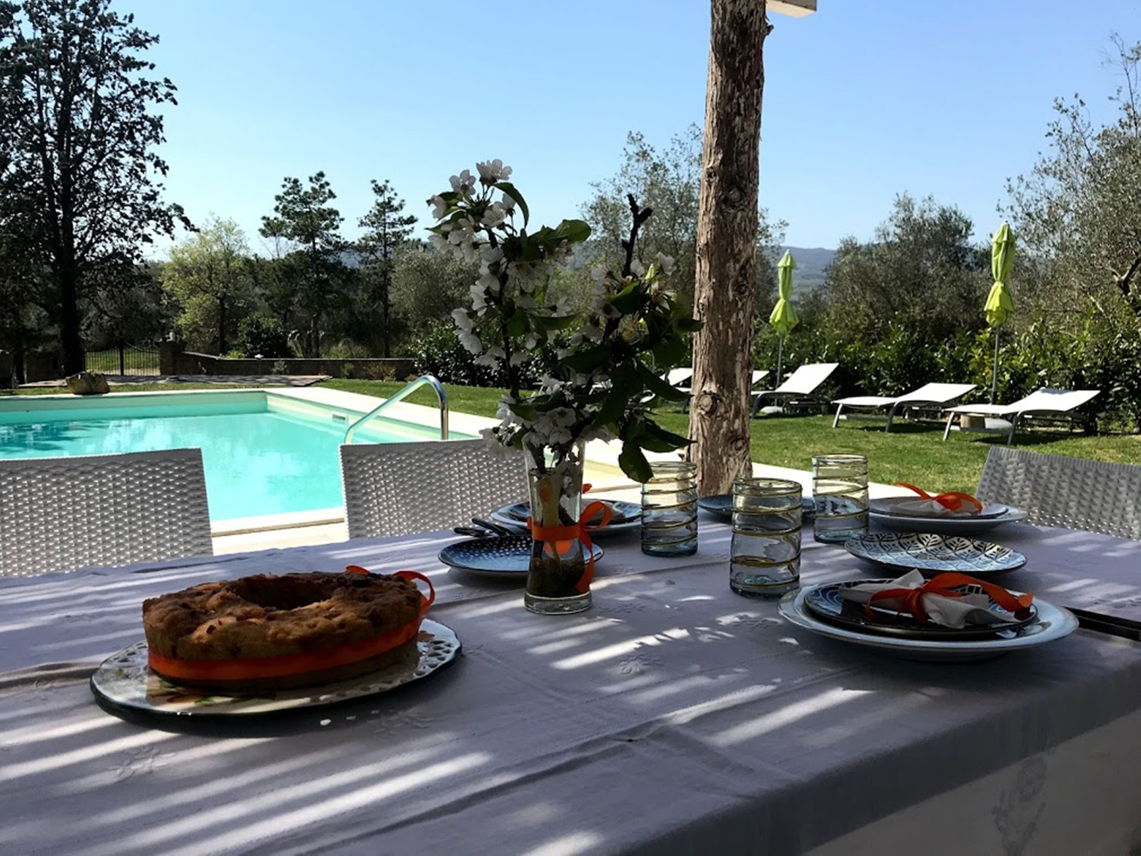 Area pranzo all'aperto con piscina sullo sfondo, tavolo apparecchiato con piatti, bicchieri e una torta.