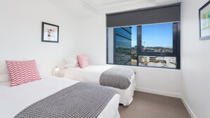 Une chambre d’hôtel moderne avec deux lits, une grande fenêtre donnant sur la ville, un ventilateur de plafond et un tableau encadré au mur.