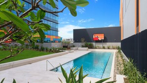 Une piscine moderne, avec un ciel bleu dégagé, entourée de verdure et bordée d’un bâtiment contemporain doté de grandes fenêtres.