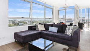 A modern living room with a sectional sofa, a glass coffee table, and large windows offering a city view. - As the Sun Sets (Zetland)
