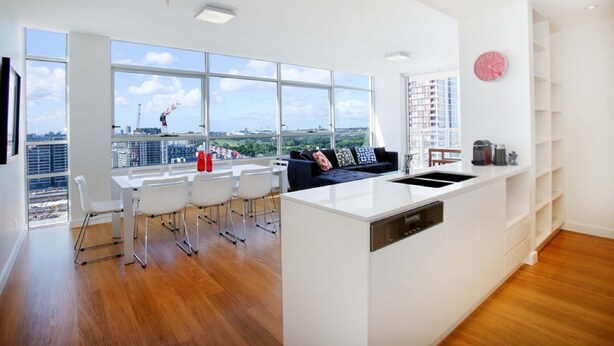 A modern kitchen with a central island, white cabinetry, and large windows offering a city view. - Moore to See (Zetland)
