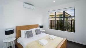 A modern hotel room with a large bed, two bedside tables with lamps, a window with a view of a fence and building, and an air conditioning unit on the wall. - Oxford Steps (Bulimba)