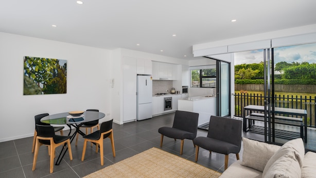 A modern kitchen with white cabinets, a dining area with a glass table and chairs, and a balcony with a view of greenery. - Oxford Steps (Bulimba)
