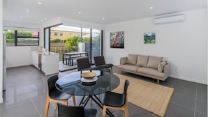 A modern living room with a glass dining table, black chairs, a beige sofa, and a large window offering a view of the outside. - Oxford Steps (Bulimba)
