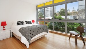 A bedroom with a large window, a bed, a nightstand, a chair, and a red lamp. - New York on Riley (Darlinghurst)